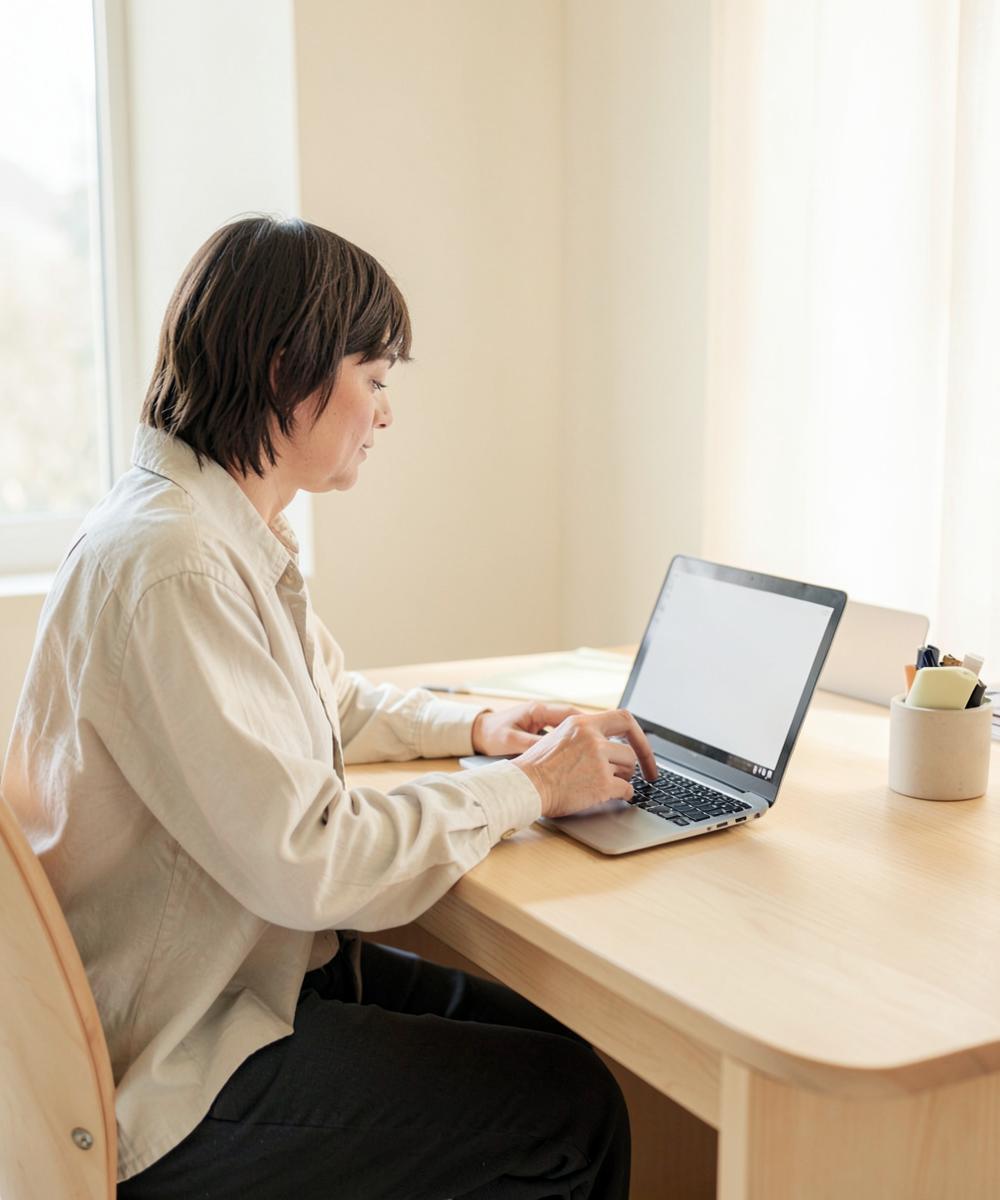 Person working on a laptop at a light oak desk in a warm minimal room